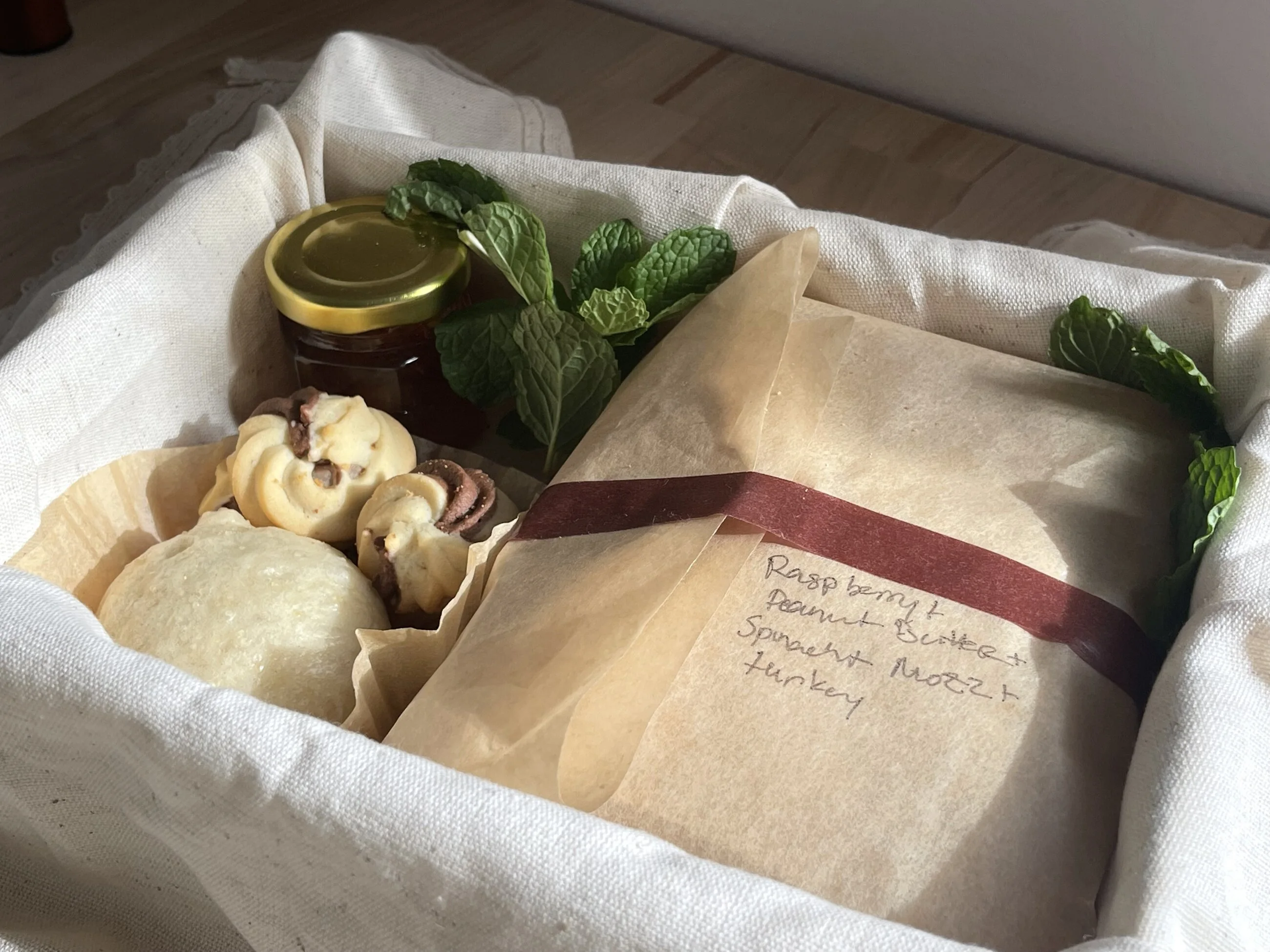 Inside the box — a jar of jam, fresh mint, butter cookies, a steamed bun, and a parcel wrapped in baking paper, sealed with a madder ribbon and labeled in pencil.