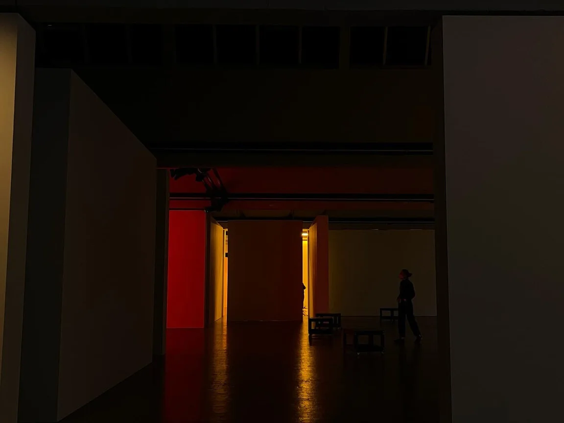 A dim gallery corridor washed in red and amber light, a lone visitor standing at its end.