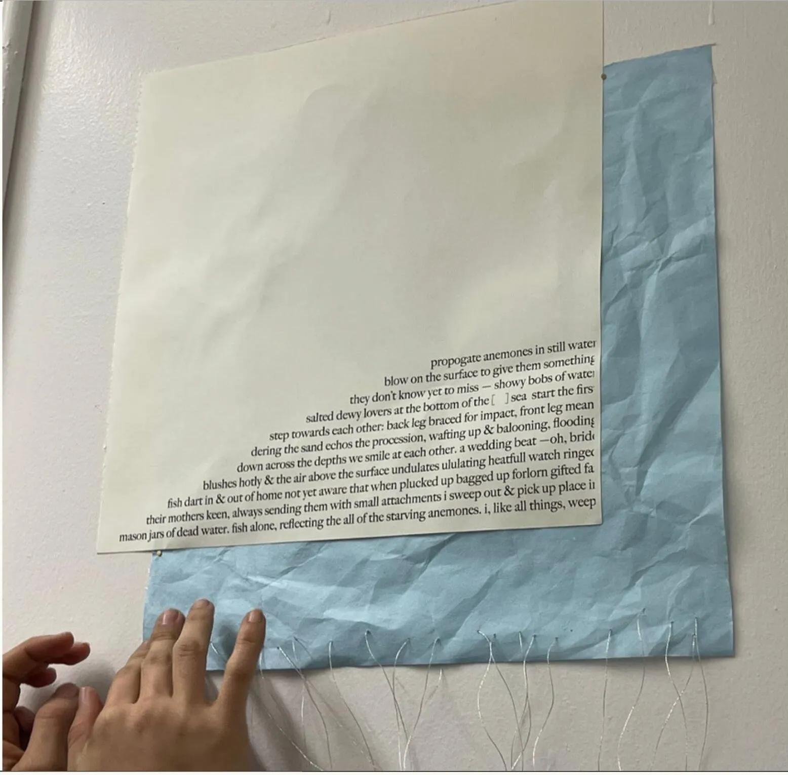 A visitor’s hands maneuver a printed sheet of the poem against crumpled blue rice paper on a gallery wall.